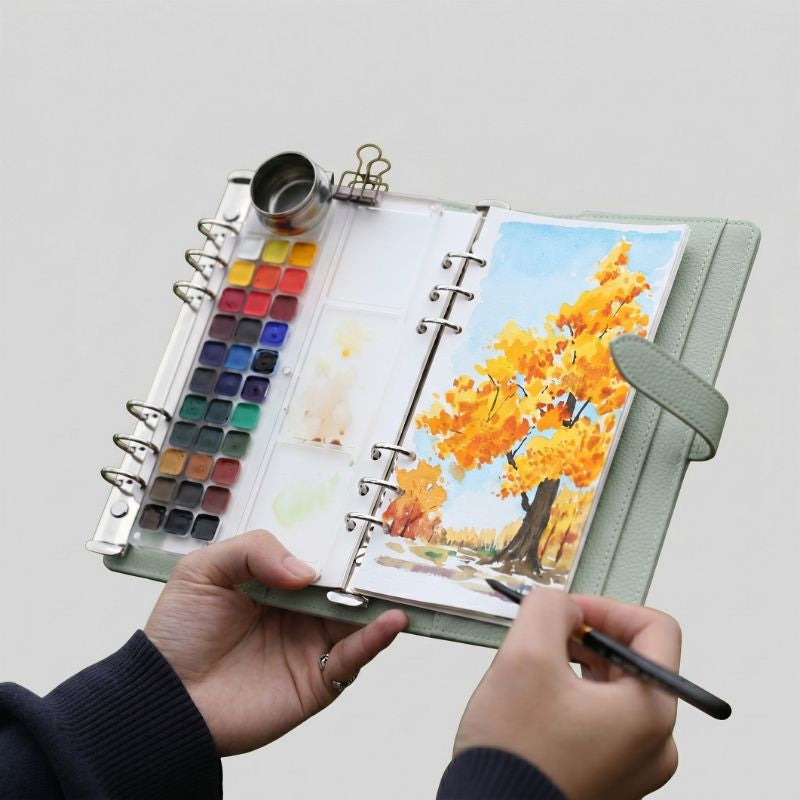 Hands holding the open Spruce Green Voyager watercolor kit, painting an autumn tree with the 33-pan palette and water pot