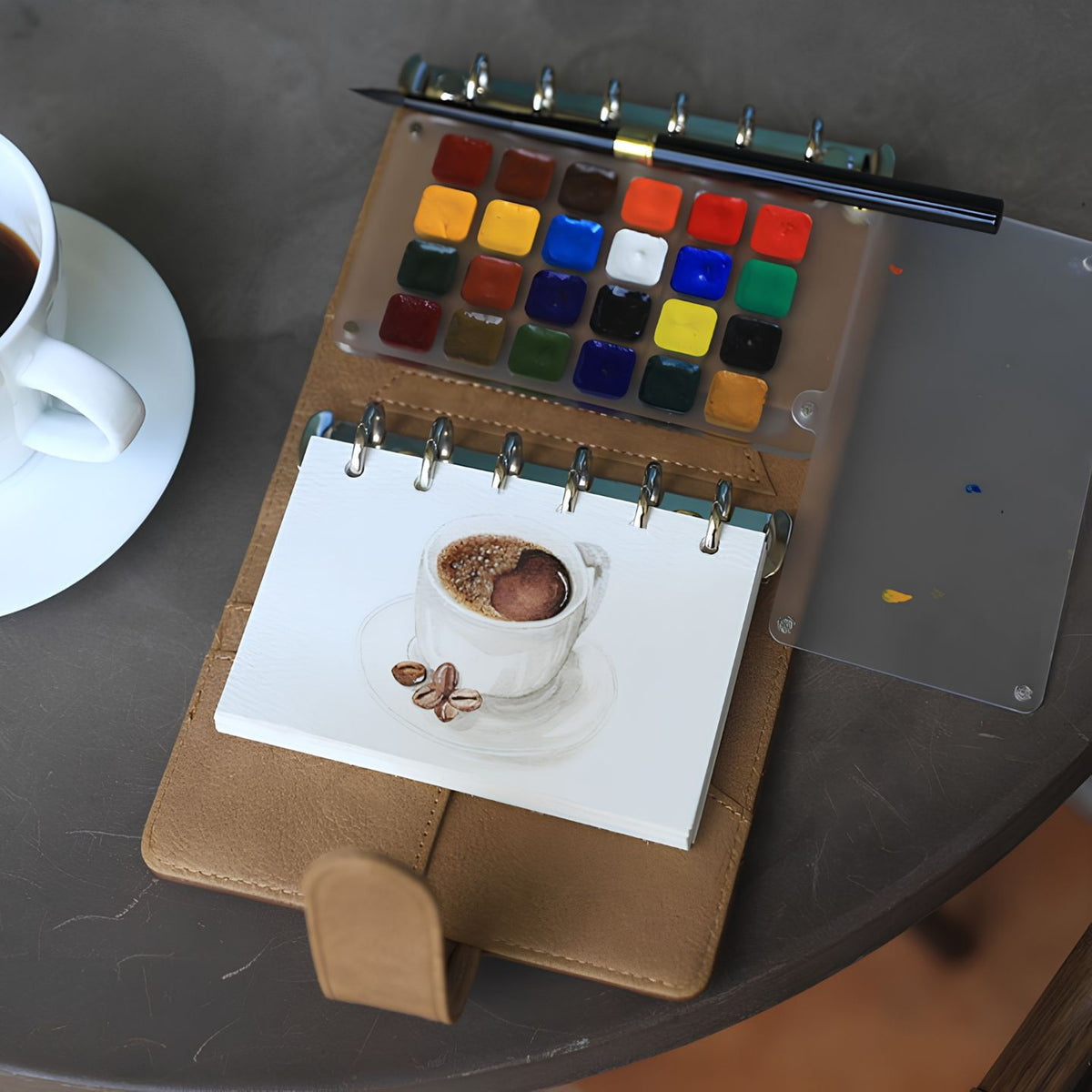 Tan leather Nomad watercolor kit open on a cafe table by an espresso, with a painted coffee-cup sketch