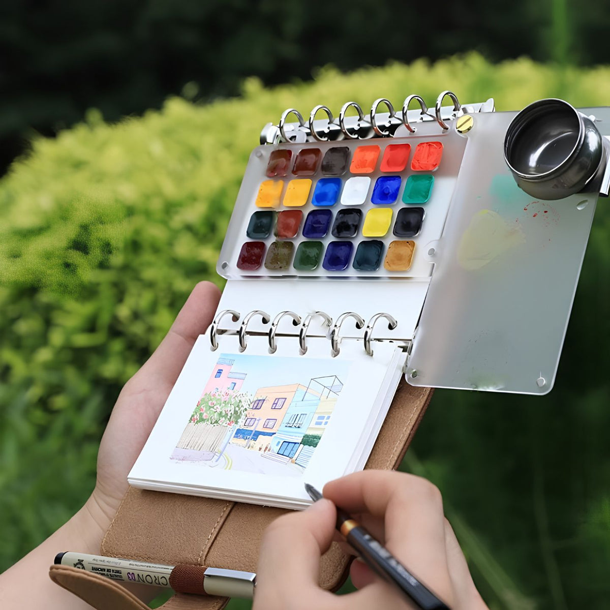 Open Nomad kit held outdoors with a 24-pan palette, water cup and a street-scene watercolor sketch