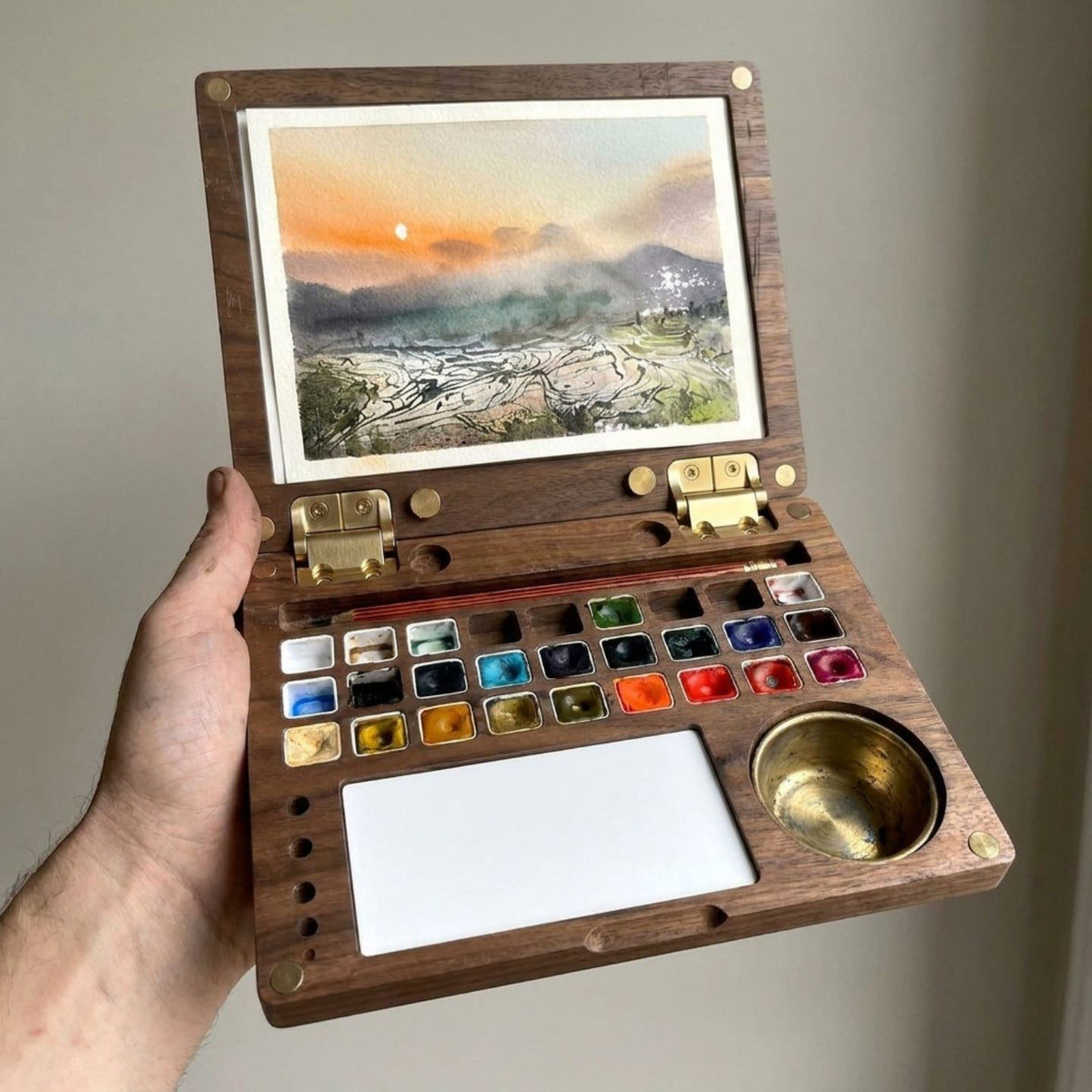 Hand holding the open ArtMate Master Edition walnut watercolor box, a finished sunset rice-terraces painting clipped to the lid above the 27-pan palette, mixing tile and brass water pot