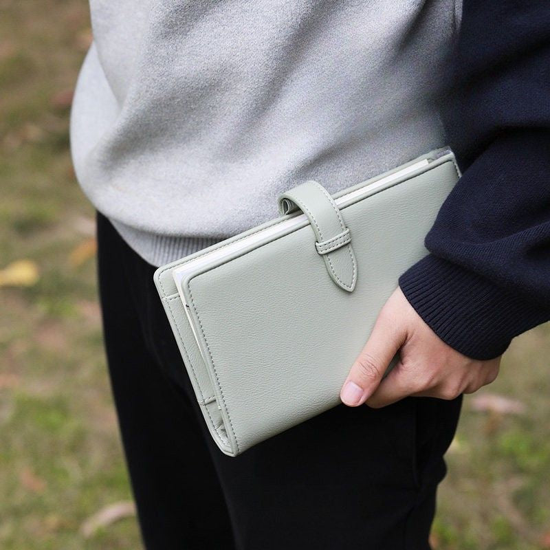 Person carrying the closed Spruce Green Voyager leather watercolor kit at their side outdoors, with the snap-tab closure and ring binder visible